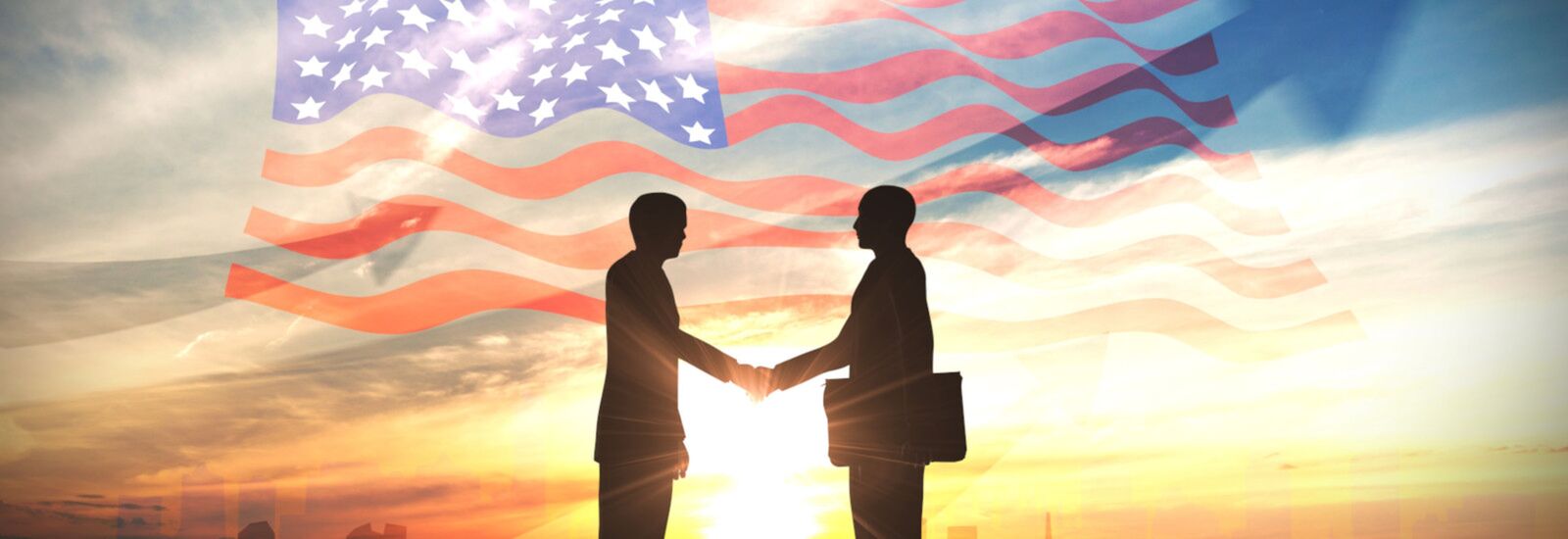 Two people shaking hands with sunset and American flag in background.